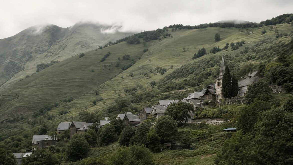 Vista panoràmica actual de Bausen, a la Val d’Aran, amb les cases de pedra i teulades de pissarra agrupades al vessant, l’església de Sant Pèir i les muntanyes cobertes de prats i boscos al fons.