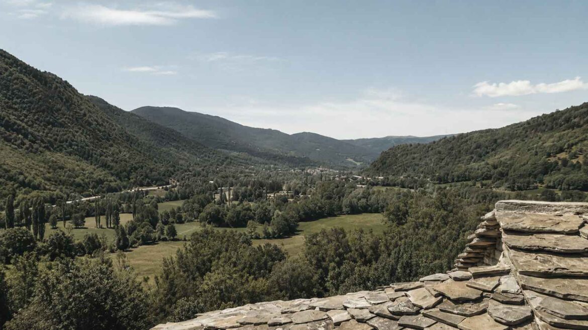 Vista de la Vall de Barravés, a l’Alta Ribagorça, amb prats al fons de vall, boscos i vessants muntanyosos coberts de vegetació.