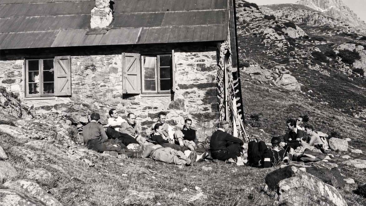 Grup d’excursionistes descansant davant del Refugi de Vallferrera, a la Vall Ferrera, l’any 1946. Els excursionistes seuen i reposen a l’exterior de l’edifici de pedra, envoltats pel paisatge d’alta muntanya.