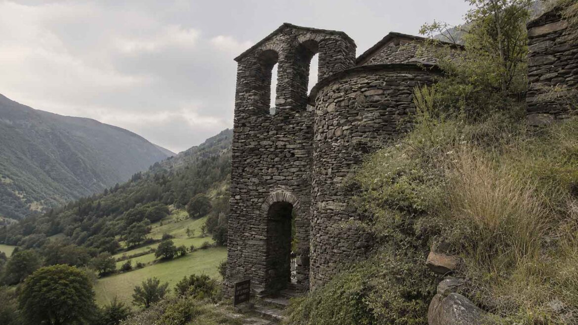 Església romànica de Sant Feliu de la Força d’Àreu, a la Vall Ferrera, amb l’absis i el campanar d’espadanya de pedra sobre els prats i vessants de la vall.