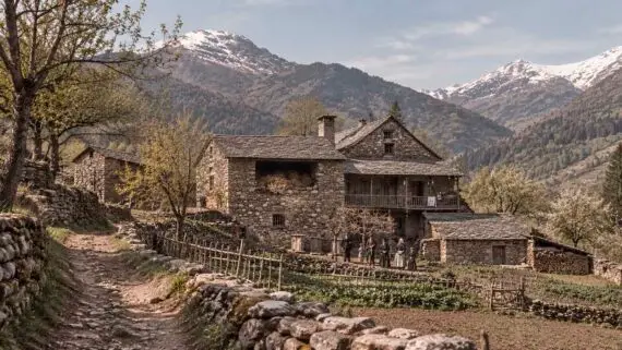 Casa pirinenca de pedra envoltada de prats, horts, camins i murs de pedra seca, amb diverses generacions de la casa davant de l’edifici i les muntanyes al fons.