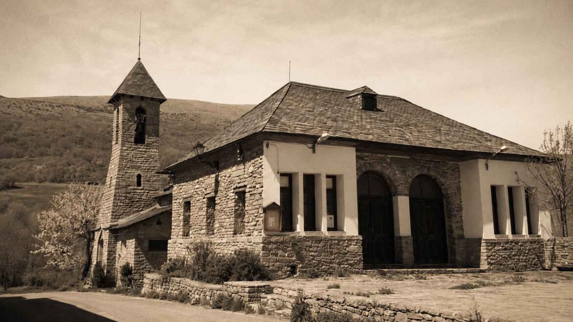 Antiga escola de Pujal, al Batlliu de Sort, al costat de l’església, en una imatge de tonalitat sèpia amb les muntanyes del Pallars al fons.